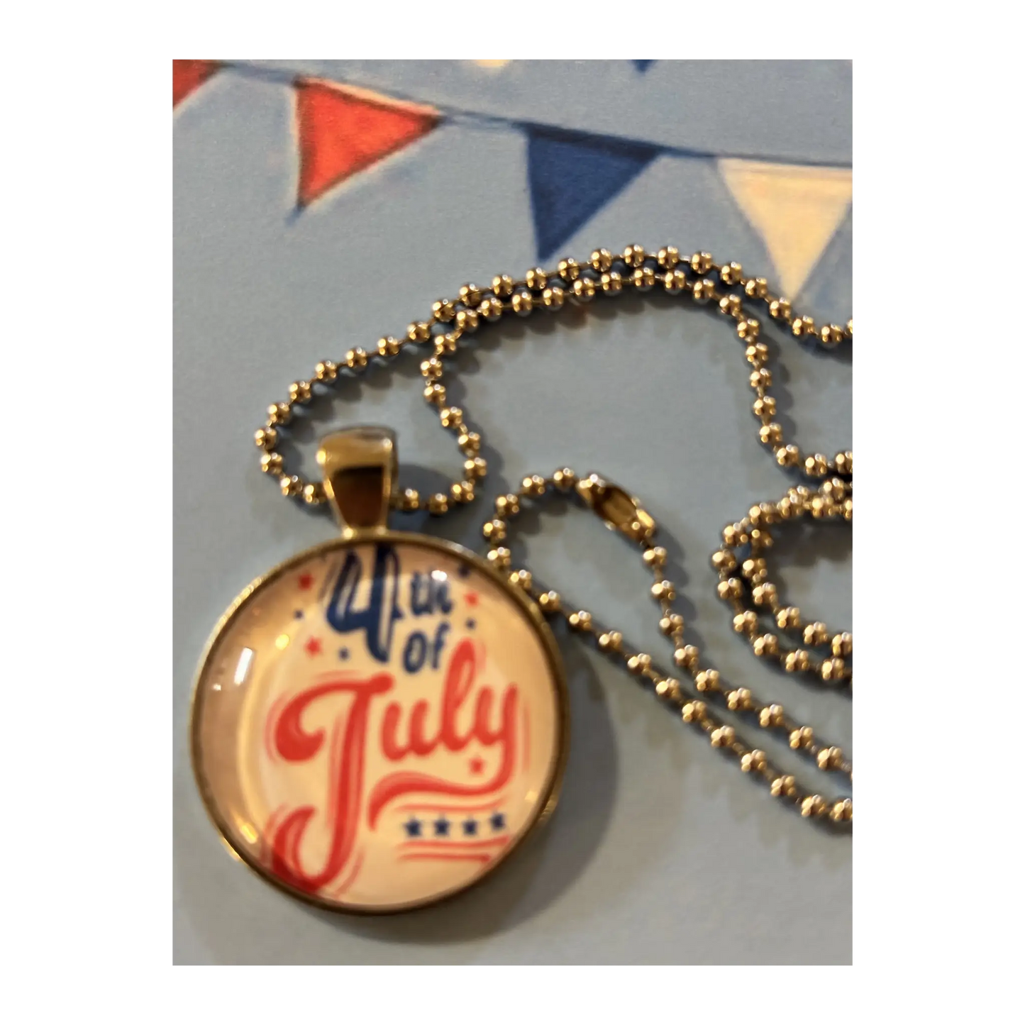 A round pendant necklace with a patriotic 4th of July design, featuring red and blue script text on a cream background under a clear dome, hanging from a silver ball chain.