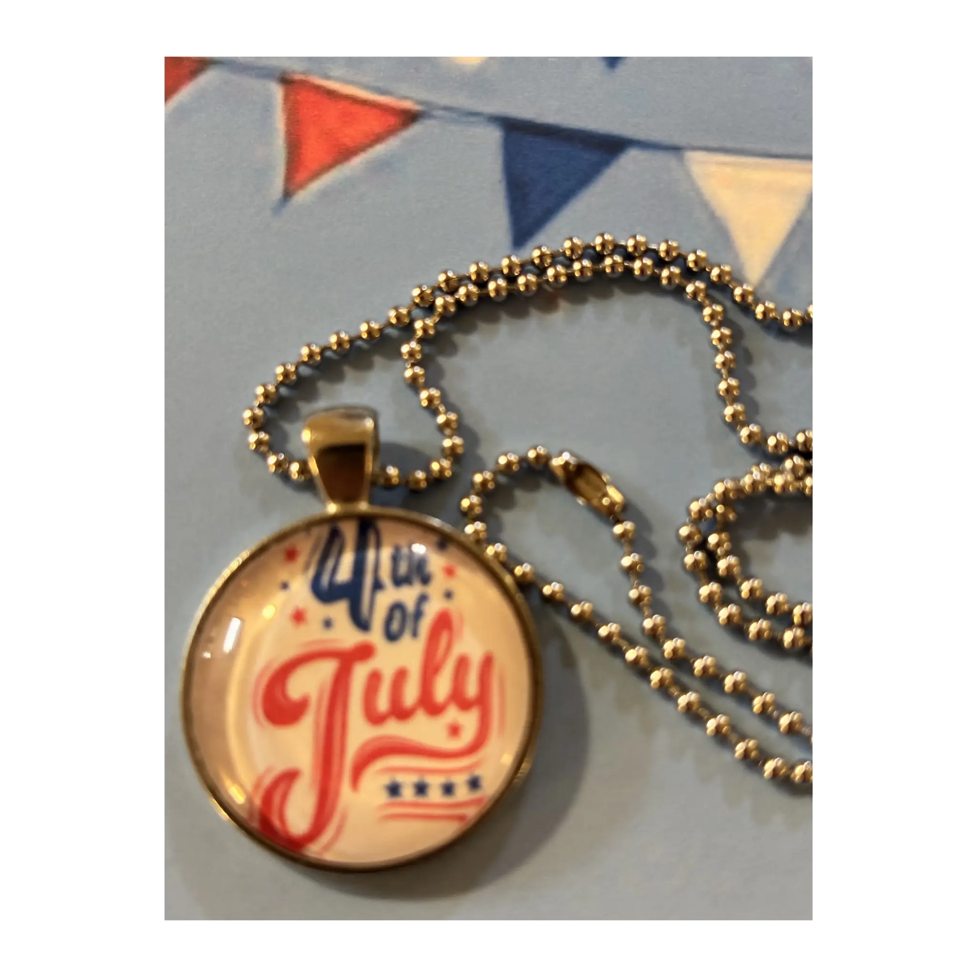 A round pendant necklace with a patriotic 4th of July design, featuring red and blue script text on a cream background under a clear dome, hanging from a silver ball chain.