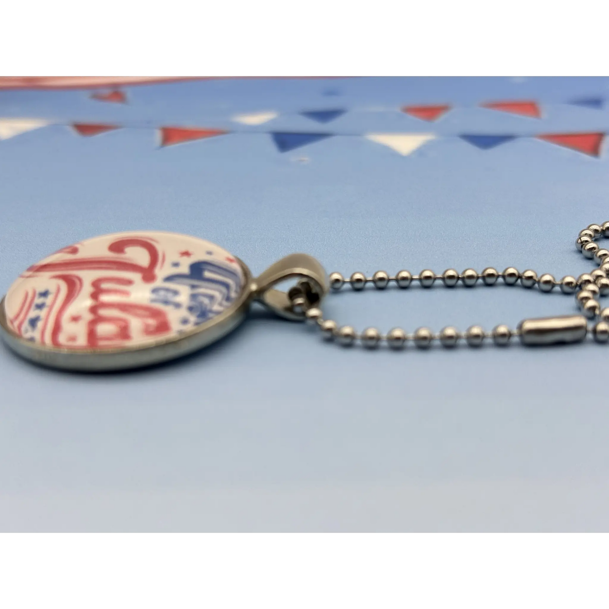 A close-up of a round pendant necklace with a patriotic red, white, and blue design, attached to a silver ball chain on a light blue background with blurred bunting.