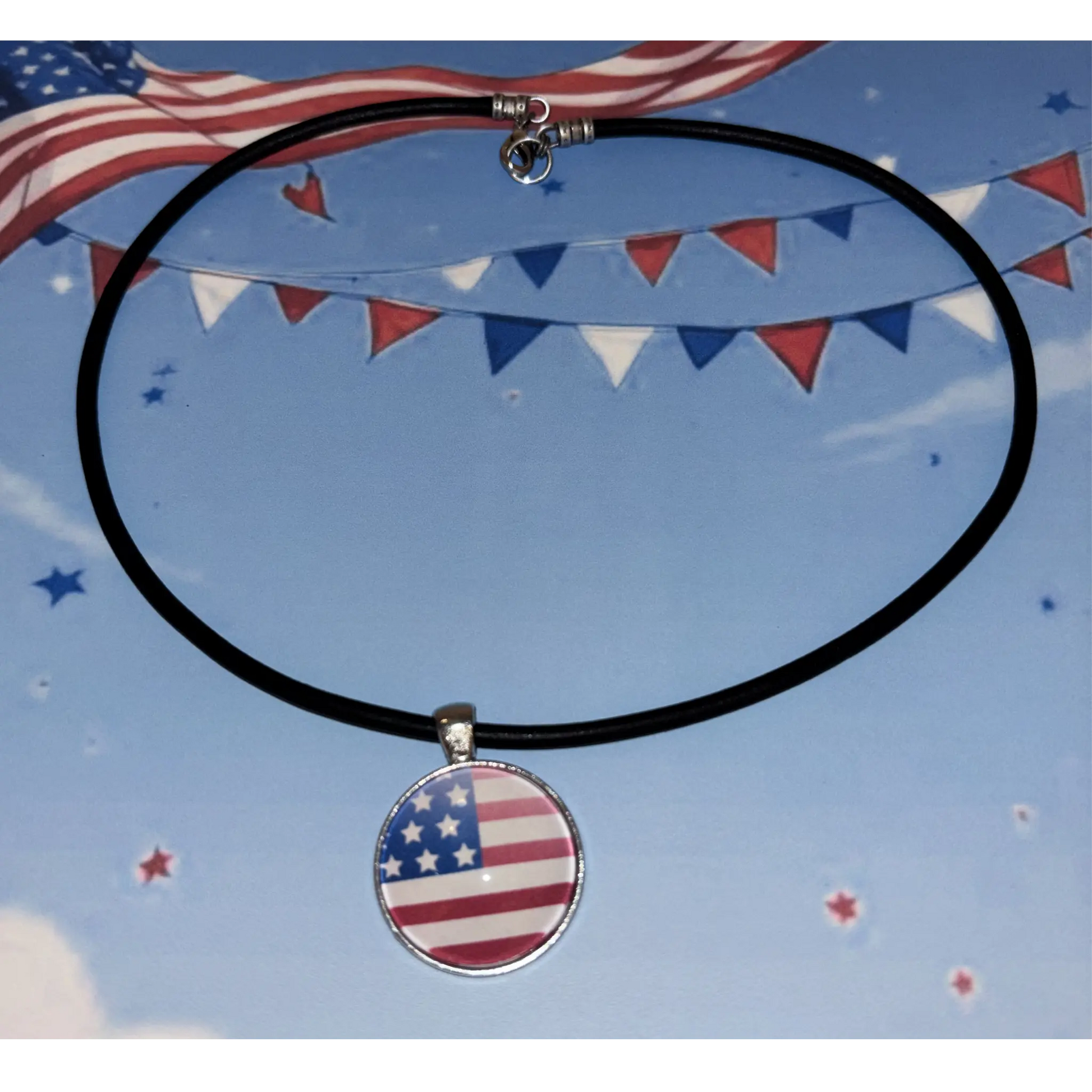 A black leather cord necklace with a round pendant featuring the American flag design, displayed against a patriotic blue sky background with bunting and stars.