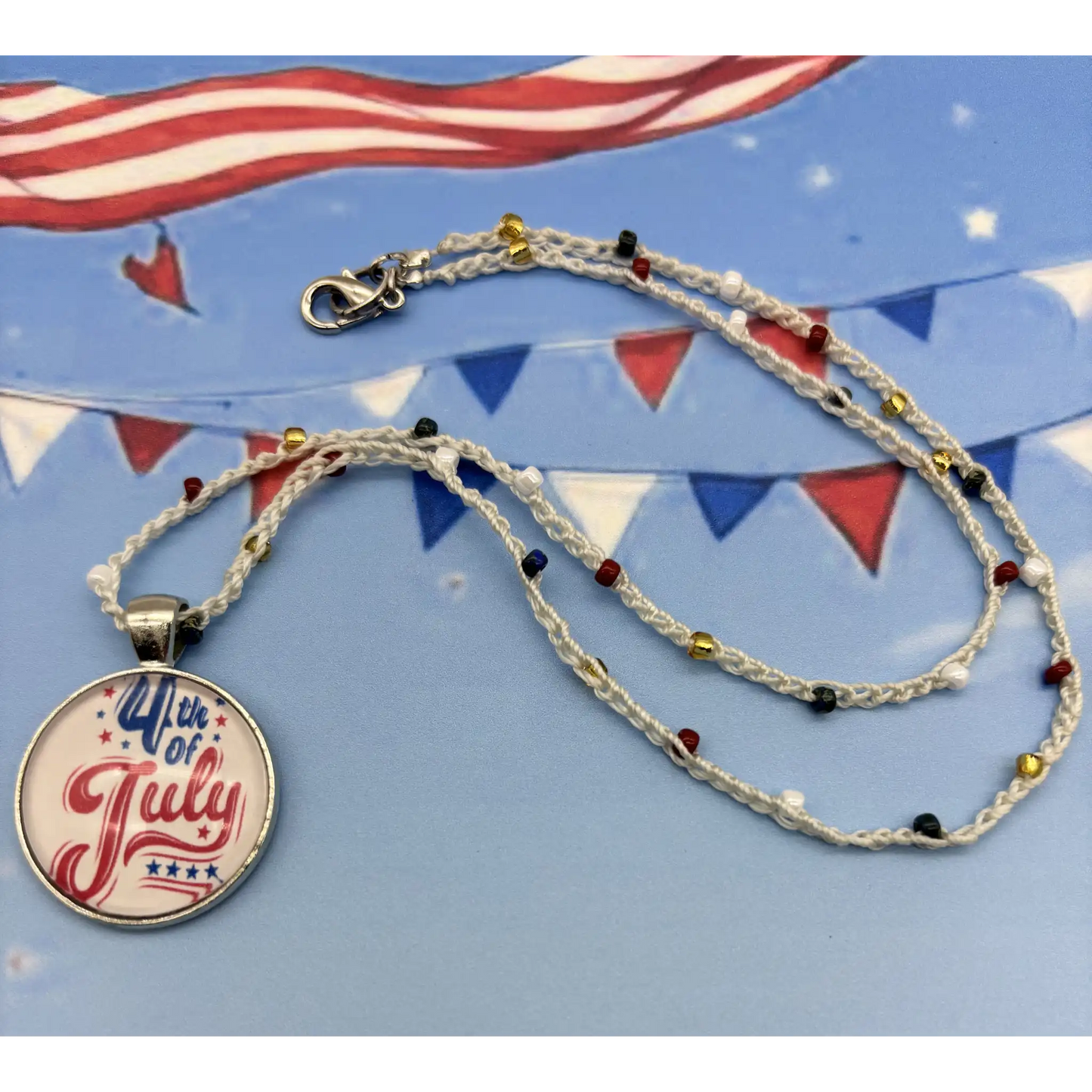 A patriotic 4th of July themed necklace featuring a round silver pendant with '4th of July' written in red and blue script on a white background. The necklace itself is made of crocheted white cord with small red, blue, white and gold beads, and is displayed on a blue background decorated with bunting flags.