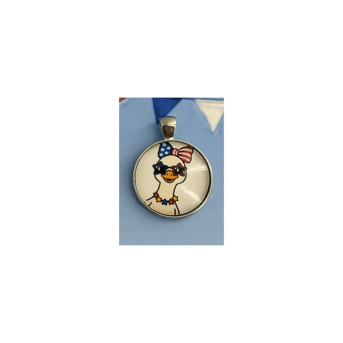 A round pendant featuring a cartoon goose wearing a red, white, and blue patriotic bow and sunglasses, set in a silver-toned bezel on a light blue background.