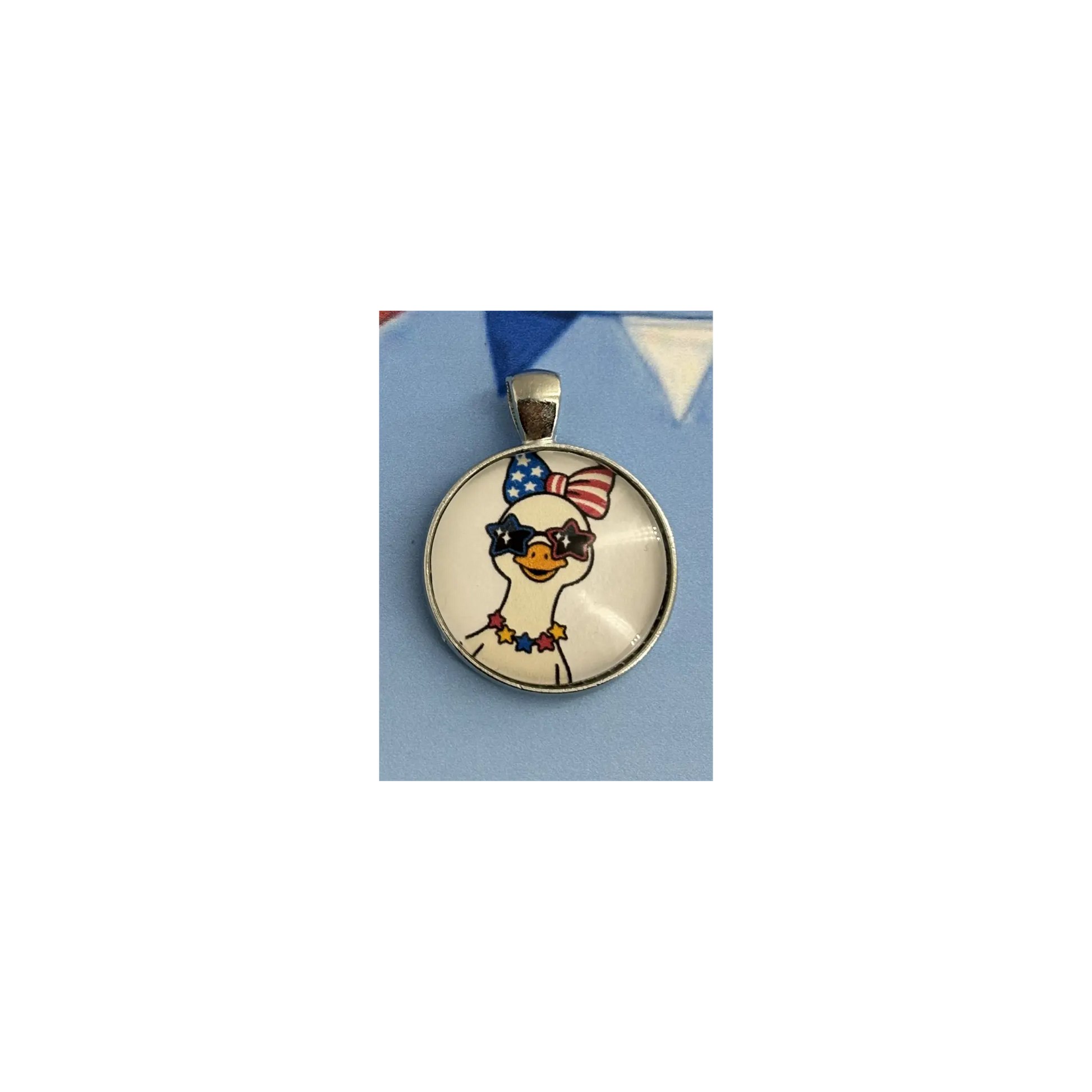 A round pendant featuring a cartoon goose wearing a red, white, and blue patriotic bow and sunglasses, set in a silver-toned bezel on a light blue background.