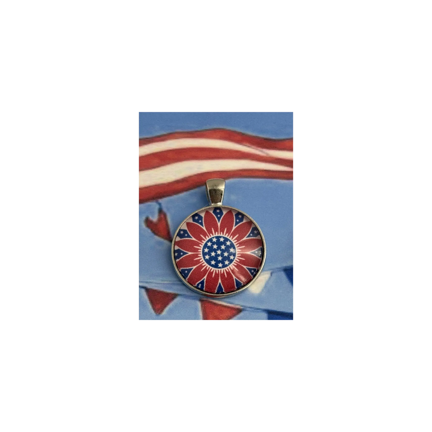 A round pendant featuring a stylized American sunflower design with red and white rays and a blue center with white stars, displayed on a blue background with a blurred American flag motif.
