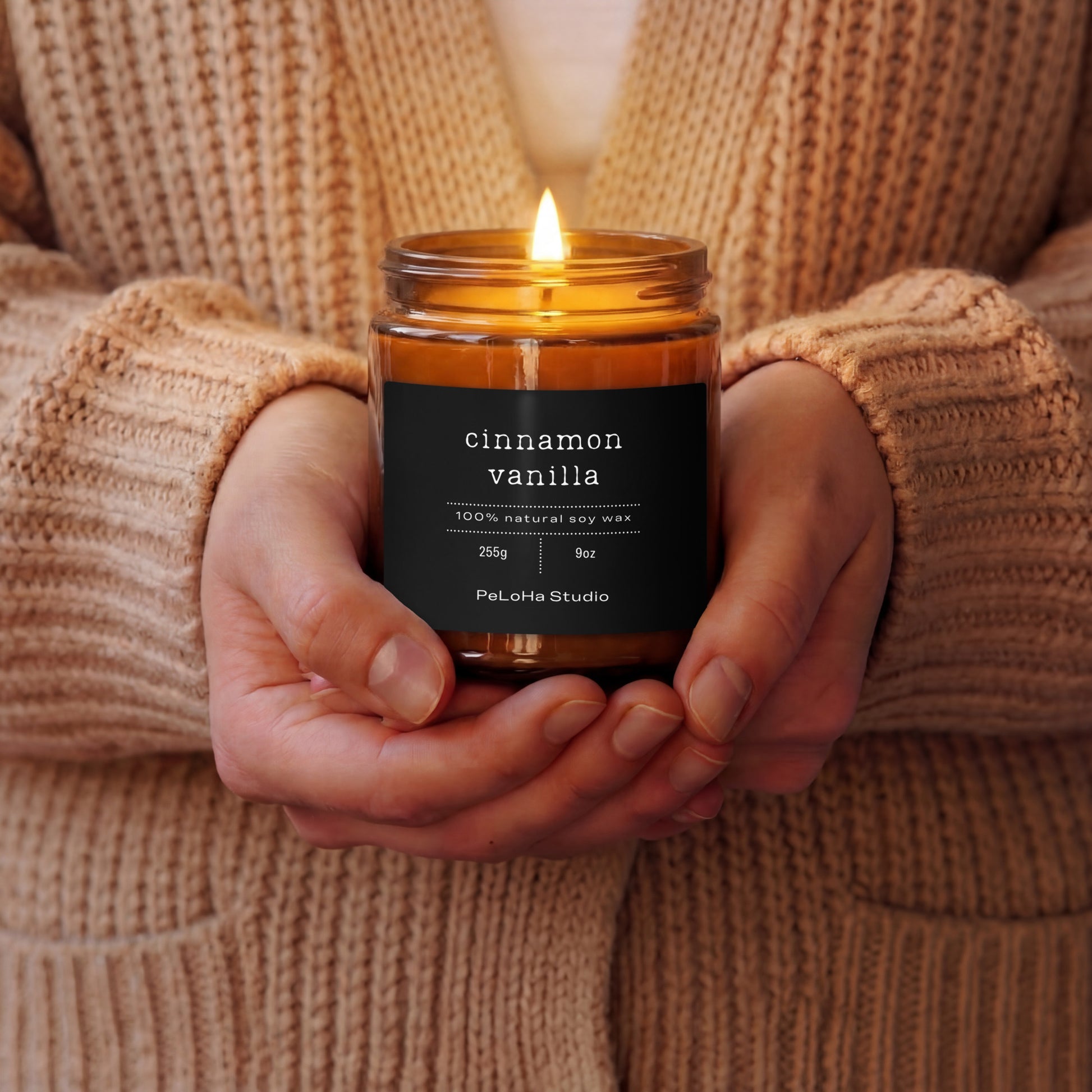 Close-up of a person cradling a glowing amber glass candle jar with a black label that reads cinnamon vanilla, 100% natural soy wax, PeLoHa Studio.