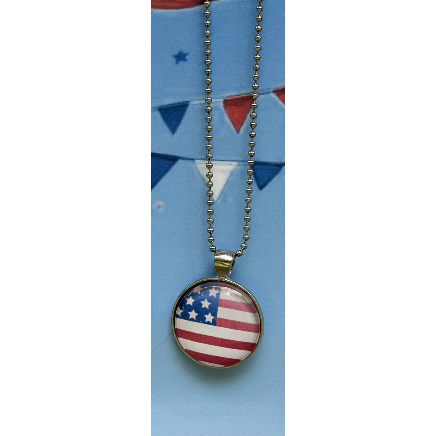 A round pendant necklace featuring the American flag design under a clear dome, hanging from a silver ball chain against a light blue background with red and white bunting.