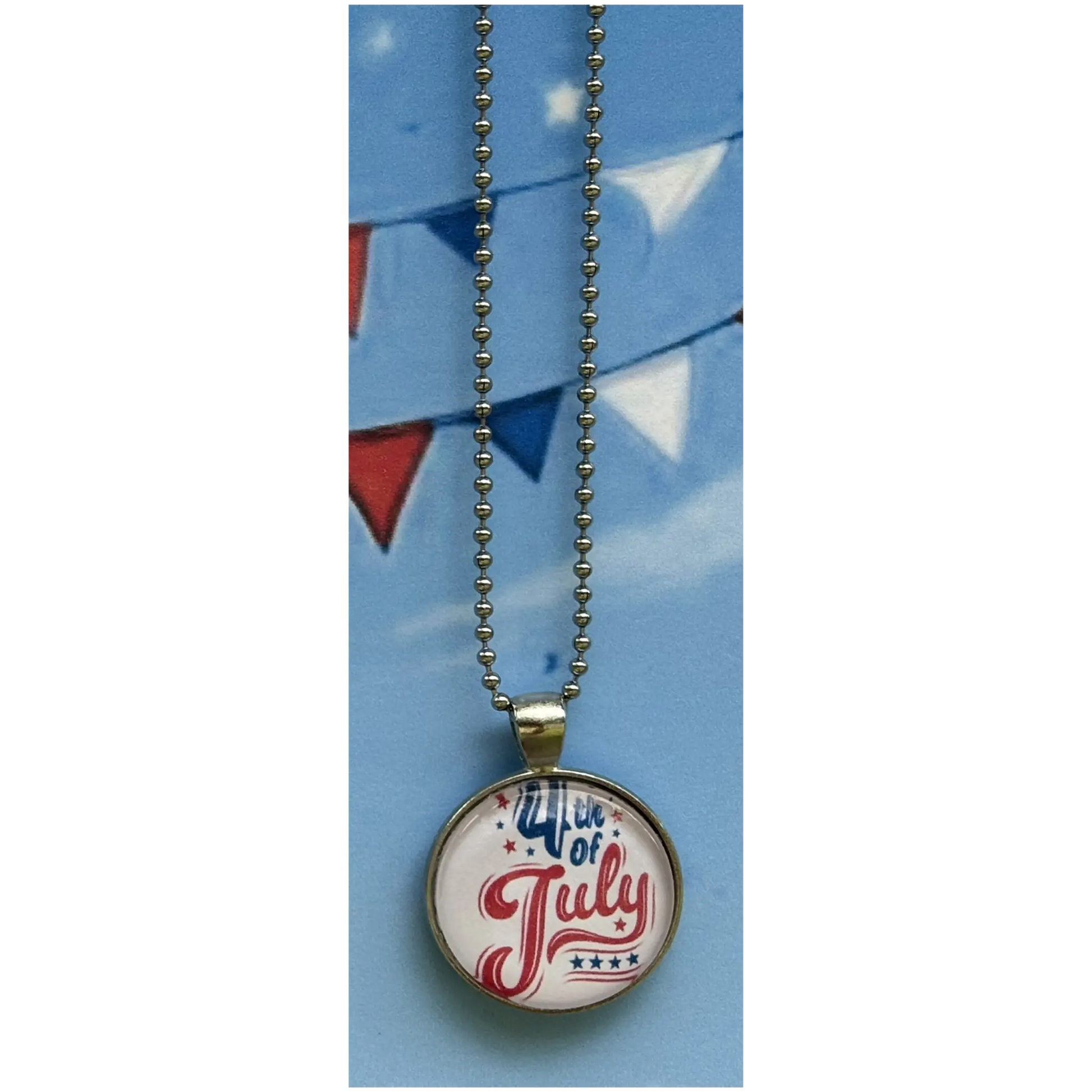 A round pendant necklace featuring a patriotic 4th of July design with red and blue script lettering on a blue background, suspended from a silver ball chain.