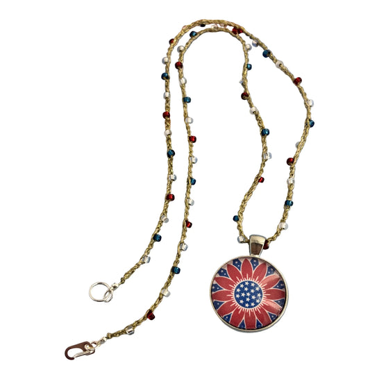 A patriotic-themed necklace featuring a round pendant with a red, white, and blue floral design, suspended on a beaded chain with small red, blue, and silver beads, displayed against a white background.