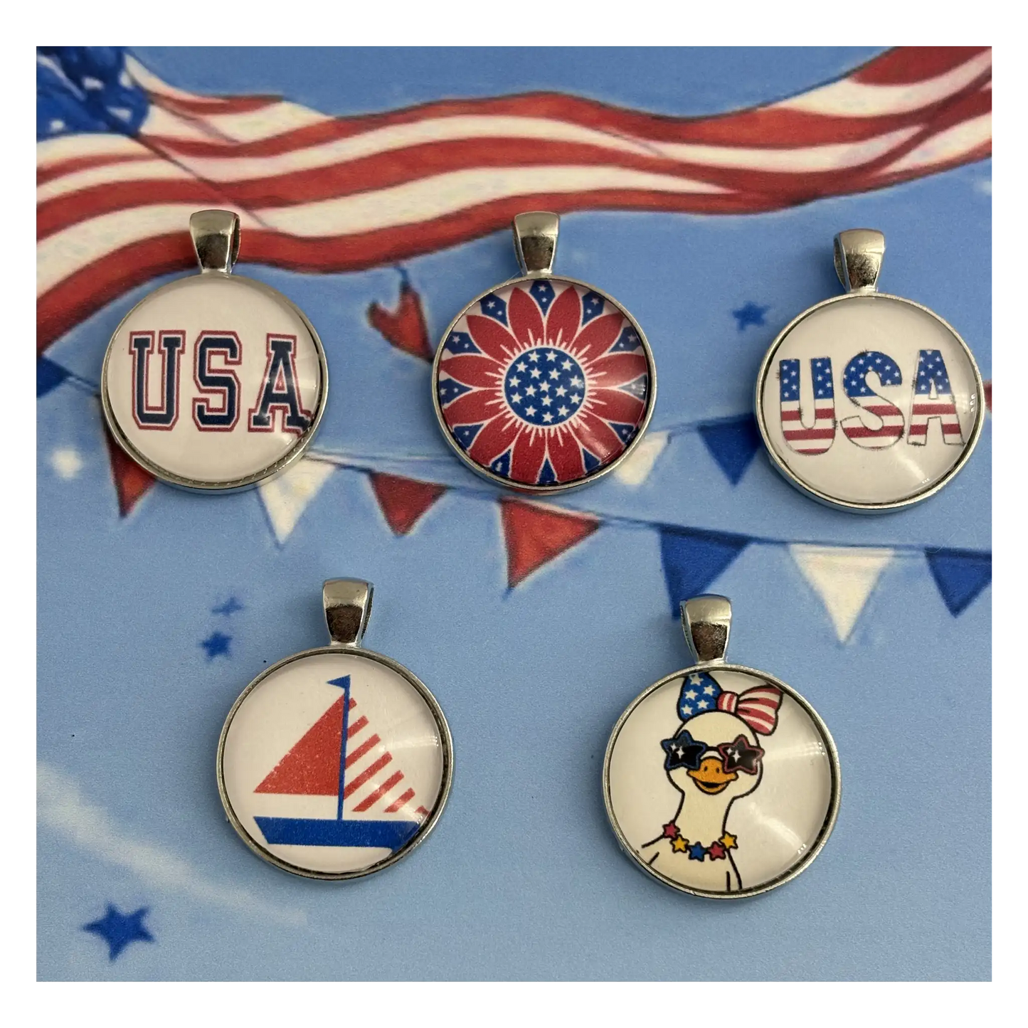 Five patriotic-themed round pendant charms with silver-toned metal rims and loops, displayed on a blue background with American flag bunting. The pendants feature designs including 'USA' in varsity lettering, a red, white, and blue sunburst, 'USA' with the American flag, a red and white sailboat, and a cartoon duck wearing sunglasses and an American flag hat.