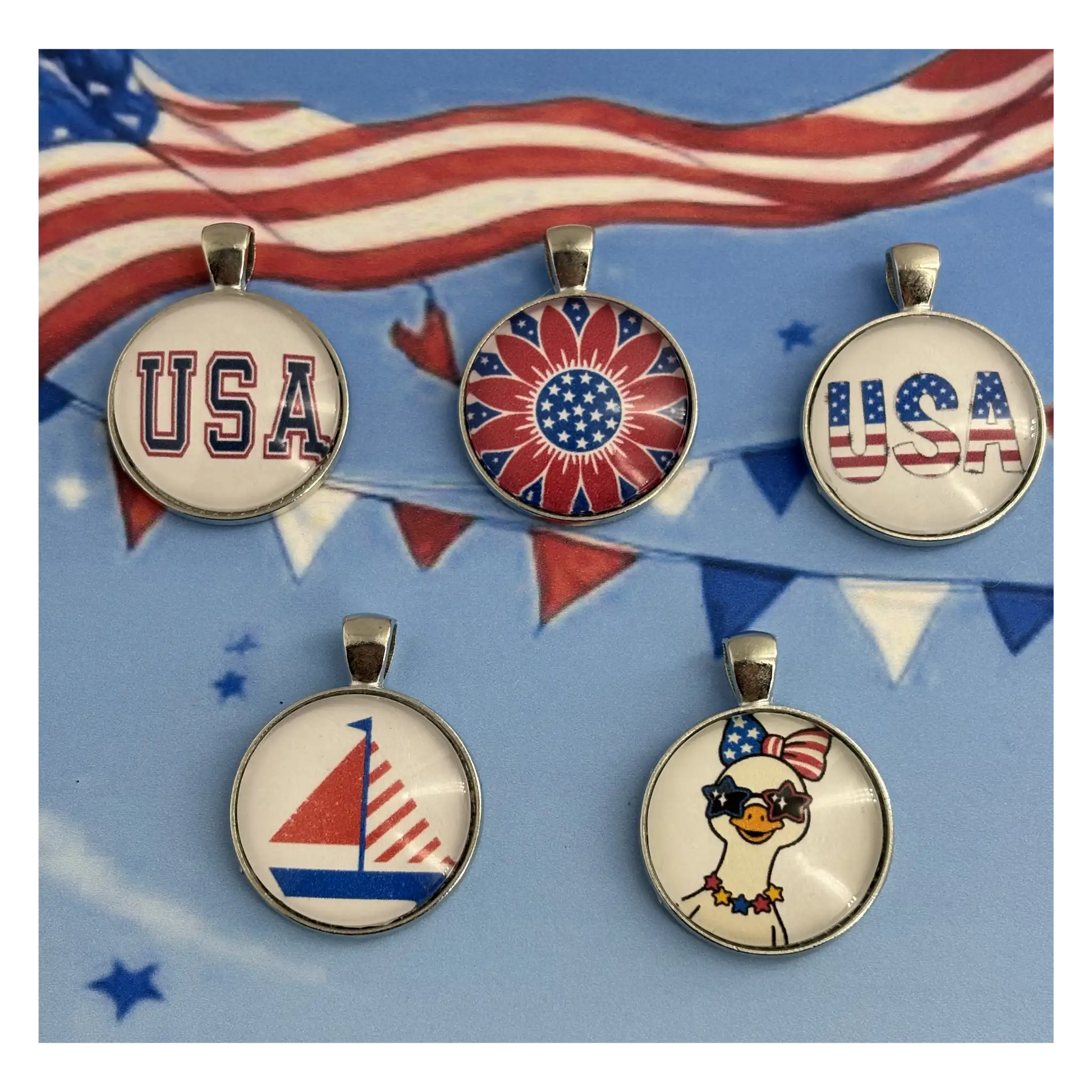 Five patriotic-themed round pendant charms with silver-toned metal rims and loops, displayed on a blue background with American flag bunting. The pendants feature designs including 'USA' in varsity lettering, a red, white, and blue sunburst, 'USA' with the American flag, a red and white sailboat, and a cartoon duck wearing sunglasses and an American flag hat.