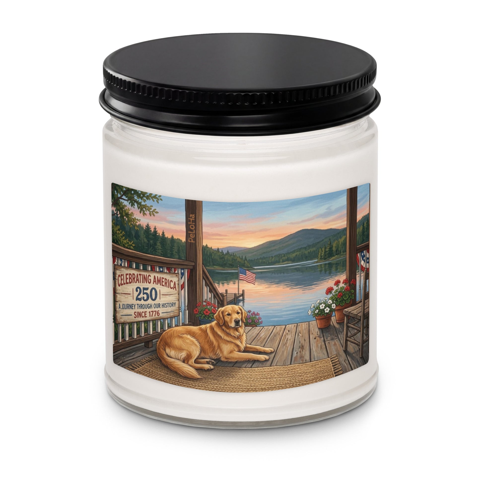 A white glass jar candle with a black metal screw-on lid, featuring a rectangular illustration on the label. The illustration depicts a golden retriever lying on a wooden deck overlooking a calm lake at sunset. A sign on the left reads "CELEBRATING AMERICA 250 | A JOURNEY THROUGH OUR HISTORY | SINCE 1776," alongside an American flag and potted flowers near the water.