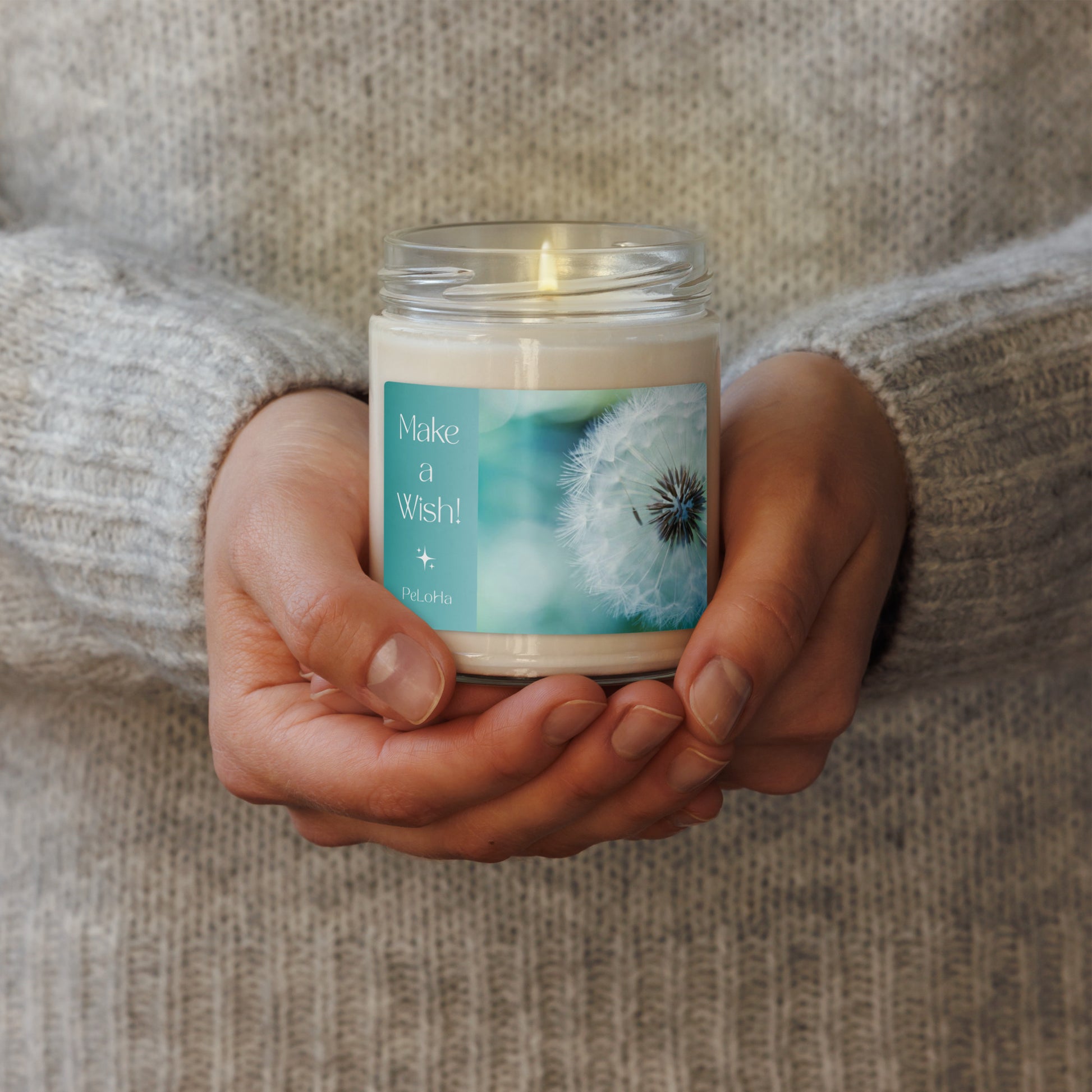 A person in a beige knit sweater holds a lit glass jar candle with a teal and white label that reads 'Make a Wish' and features a dandelion design.