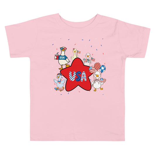A pink short-sleeve jersey t-shirt features a fun, festive print of patriotic ducks, twinkling stars, and exploding confetti on a white background.