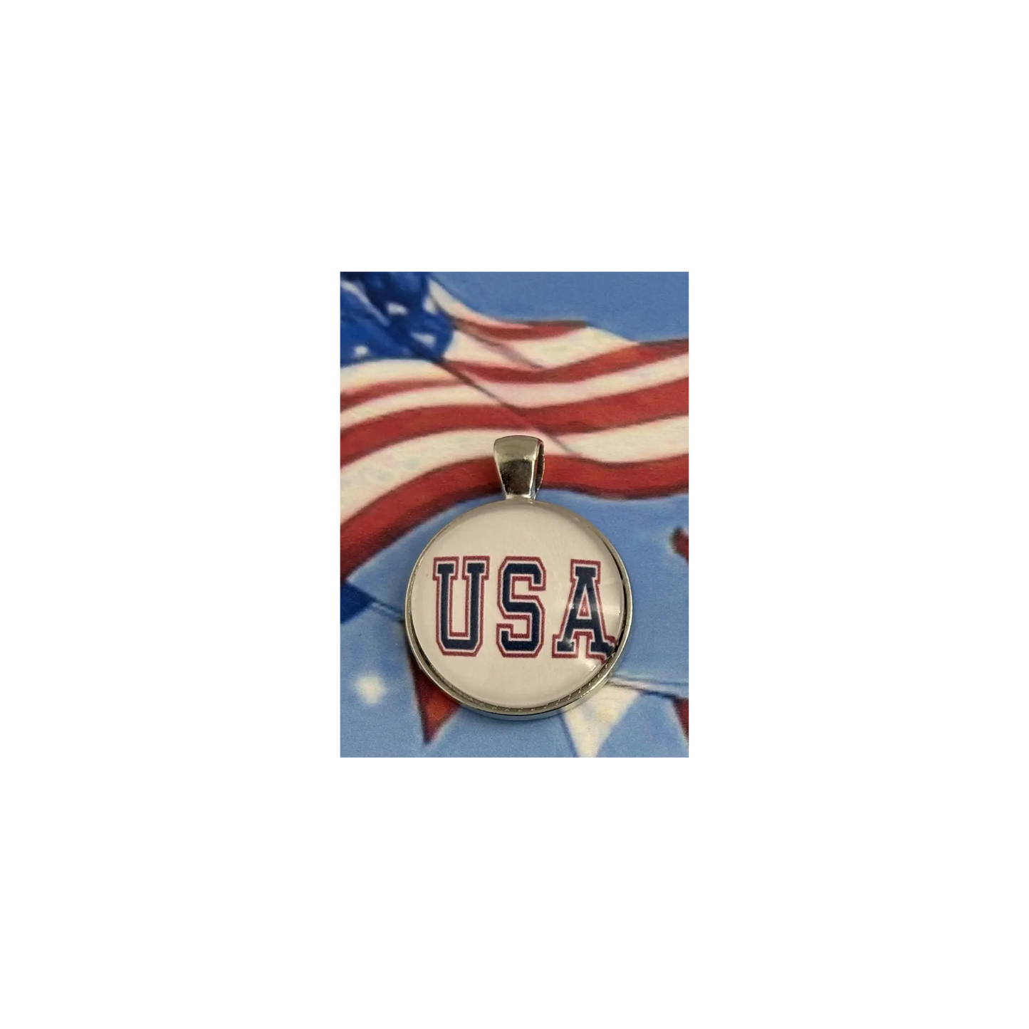 A round pendant charm featuring the letters 'USA' in navy blue with a red outline on a white background, suspended from a silver-toned bail. The pendant is centered against a background graphic of a waving American flag.