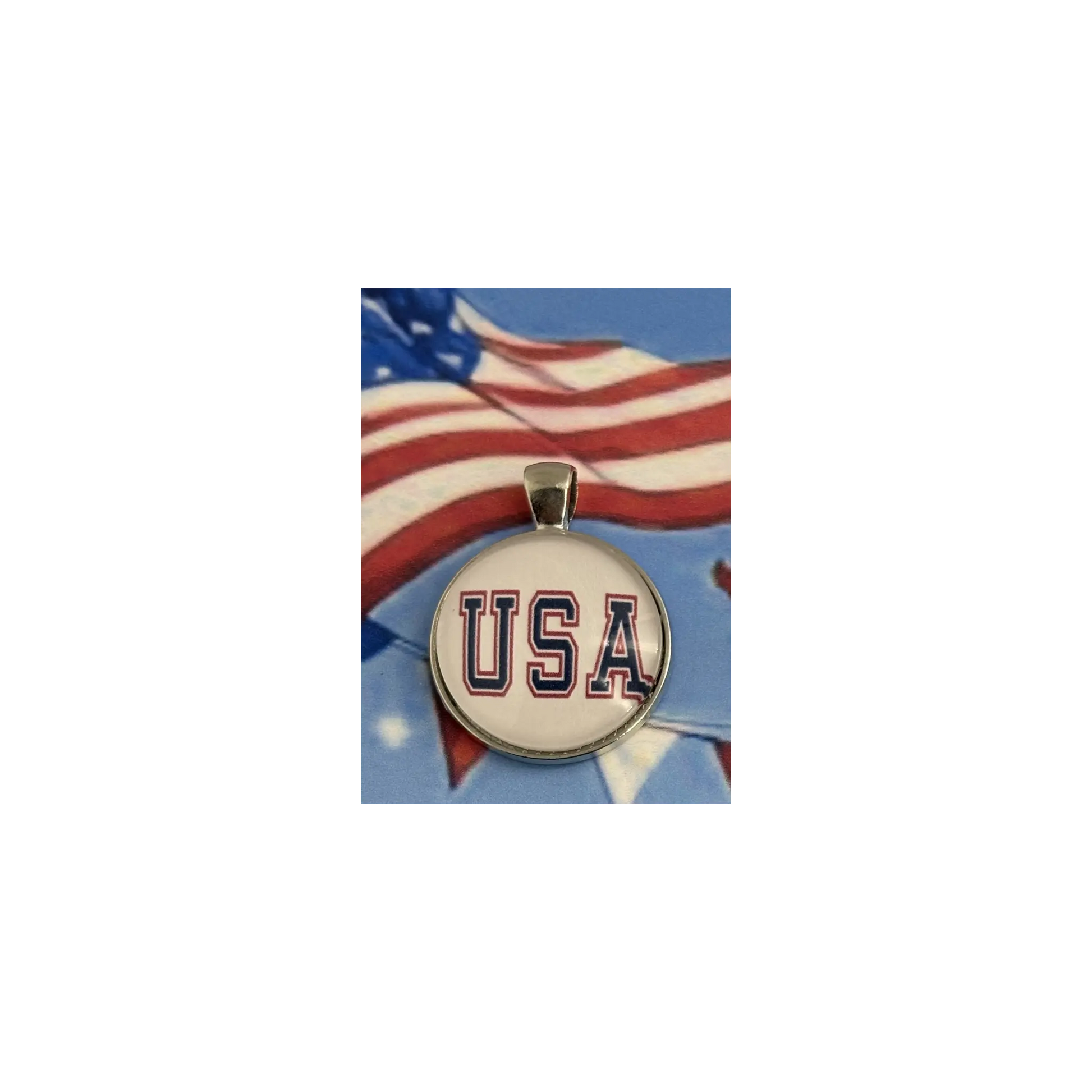 A round pendant charm featuring the letters 'USA' in navy blue with a red outline on a white background, suspended from a silver-toned bail. The pendant is centered against a background graphic of a waving American flag.