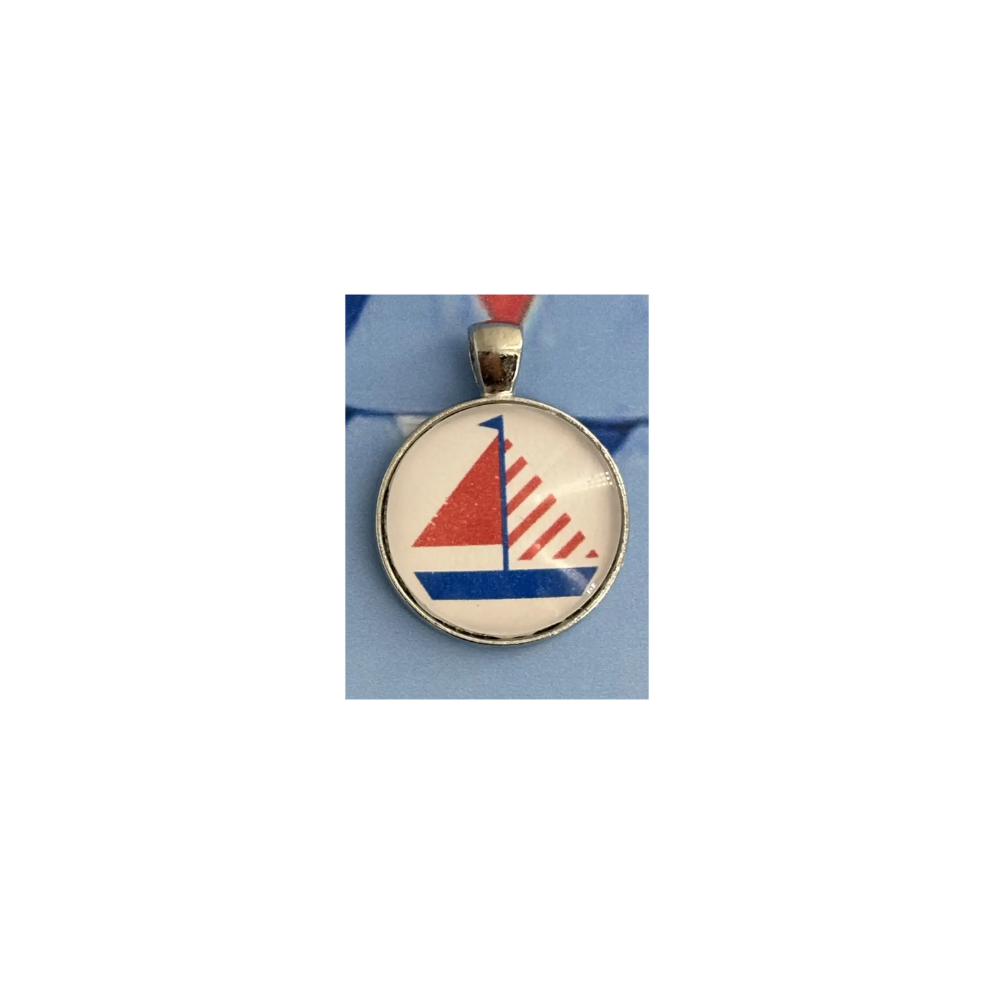 A round pendant charm featuring a stylized red, white, and blue sailboat design under a clear dome, set in a silver-toned bezel with a red ribbon loop at the top, shown on a light blue background.