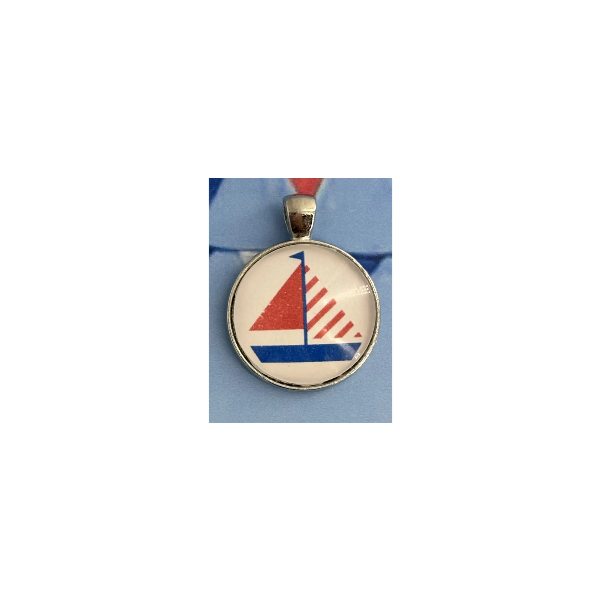 A round pendant charm featuring a stylized red, white, and blue sailboat design under a clear dome, set in a silver-toned bezel with a red ribbon loop at the top, shown on a light blue background.